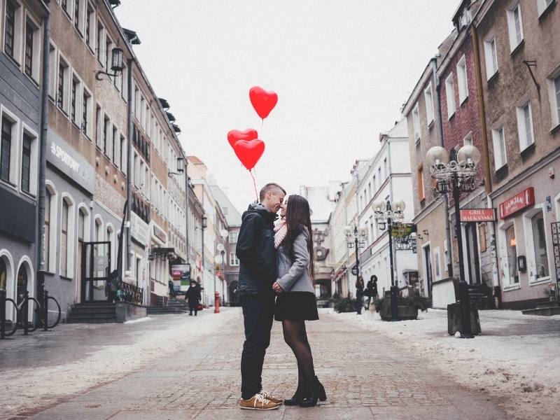 man kissing woman below heart balloons